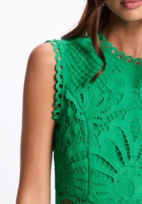 Top en dentelle verte avec des motifs floraux, des bords festonnés et des découpes délicates. Le matériau semble léger avec une surface texturée.