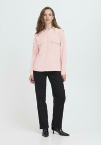 Helles rosa Langarmhemd mit Knopfverschluss, zwei Brusttaschen, kombiniert mit schwarzen Hosen und spitzen schwarzen Absätzen. Glatte Stofftextur.