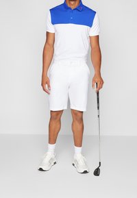Herrgolfoutfit med en blå och vit polo, vita shorts och vita sneakers med svarta detaljer, som håller en golfklubba.