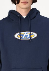 Sweat à capuche bleu marine en coton, doté d'un grand logo brodé en bleu et jaune. Comprend une poche avant et des cordons de serrage ajustables.