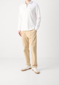 Mężczyzna ubrany w białą koszulkę polo z długim rękawem, beżowe spodnie i białe sneakersy z gumowymi podeszwami, stojący na jednolitym jasnym tle.