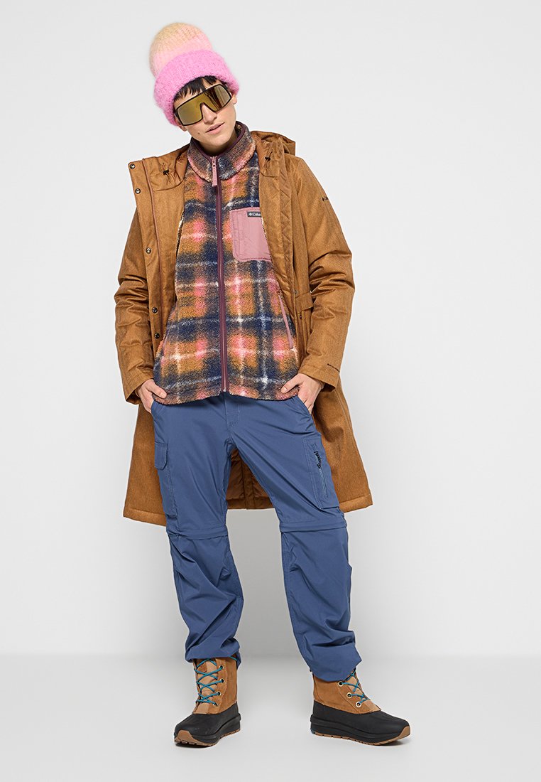Brauner wasserdichter Mantel, karierte Fleecejacke mit pinken Akzenten, blaue Outdoor-Hose und braune Stiefel mit blauen Schnürsenkeln. Pinke Strickmütze und Sonnenbrille.