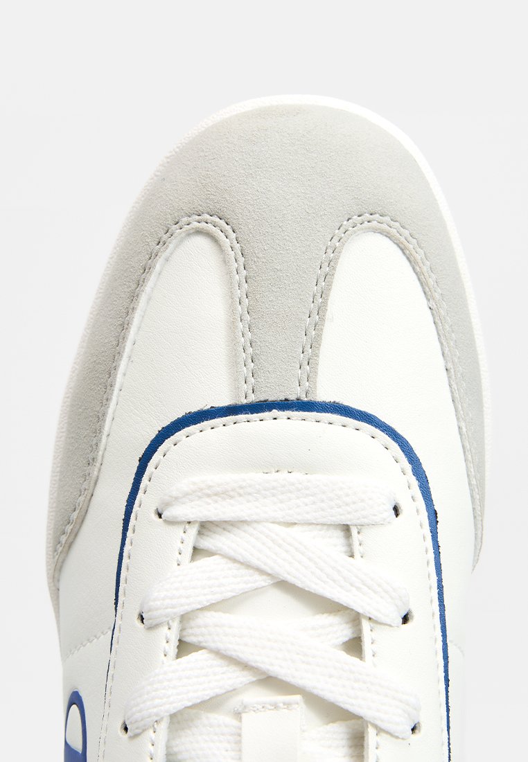 Sneaker bianco con accenti in suede grigio, dettagli blu, punta rotonda e lacci bianchi. Design testurizzato con bordi cuciti e logo sul lato.