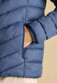 Marineblå pufferjakke med chevron-syninger, der har en lynlåslomme og elastiske manchetter. Blød, glat tekstur med et mærke-logo.