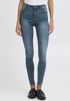 Frau trägt hoch taillierte enge blaue Jeans kombiniert mit schwarzen Loafern und steht vor einem schlichten weißen Hintergrund.