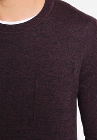 Dunkelburgunderfarbener Strickpullover mit Rundhalsausschnitt, strukturiertem Gewebe und einer kleinen Brusttasche. Hergestellt aus weichem, warmem Material.