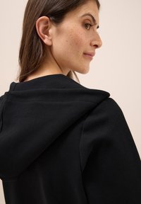 Felpa nera con cappuccio e texture a costine. Presenta un grande cappuccio e dettagli di cucitura visibili. Il tessuto appare morbido e informale.