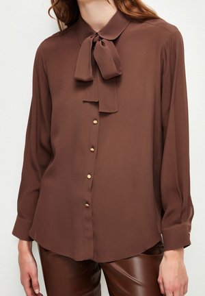 Bruine blouse met lange mouwen, gouden knopen en een gestrikte strik bij de hals, gedragen met bruine leren broeken, getoond van midden op de borst tot aan de heupen.