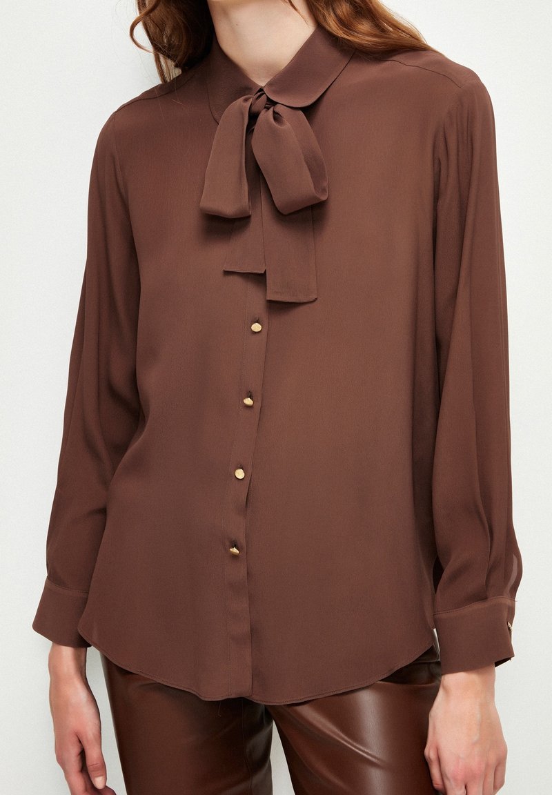 Blouse marron à manches longues avec des boutons dorés et un nœud noué au cou, portée avec un pantalon en cuir marron, montrée de la moitié de la poitrine aux hanches.