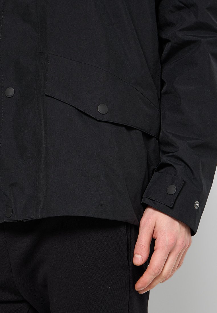 Zwarte jas met drukknopen-flapzak en manchetdetail op de linkerarm, gedragen door een persoon met een zichtbare rechterhand en zwarte broek.
