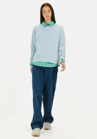 Jasnoniebieski, długi rękaw, bluza z zaokrąglonym dołem, noszona na zielonej koszuli z kołnierzykiem, w połączeniu z luźnymi, ciemnoniebieskimi dżinsami i jasnymi sneakersami.