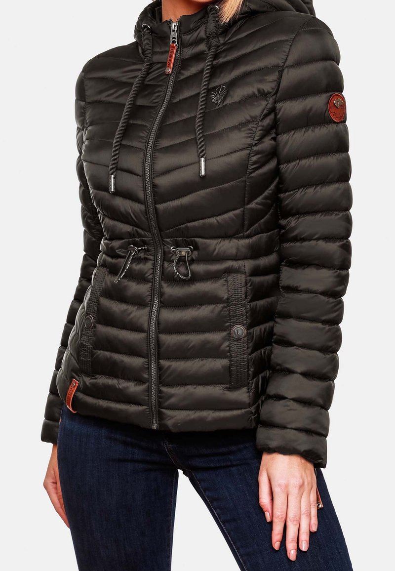 Femme portant une veste noire matelassée cintrée à capuche avec fermeture éclair, taille à cordon et deux poches avant sur un jean bleu foncé.