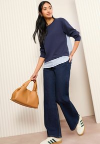 Felpa navy con orlo a righe, abbinata a jeans a gamba larga scuri. Borsa beige in mano, sneakers bianche e verdi ai piedi.