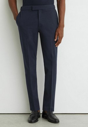 Mann trägt marineblaue Slim-Fit-Herrenhose und schwarze Lederloafer, steht mit einer Hand in der Tasche vor einer weißen Wand.