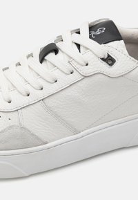 Zapatillas de cuero blancas con paneles laterales texturizados, acentos de ante gris y una lengüeta negra con logo. Diseño estándar de cordones.