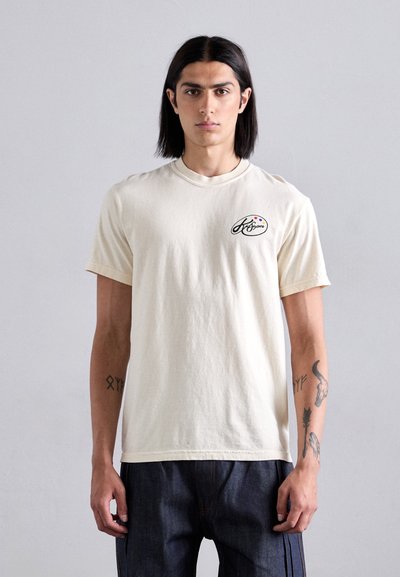 Cream-colored cotton t-shirt with a crew neckline, short sleeves, and a small logo detail on the left chest. Smooth texture, casual fit.