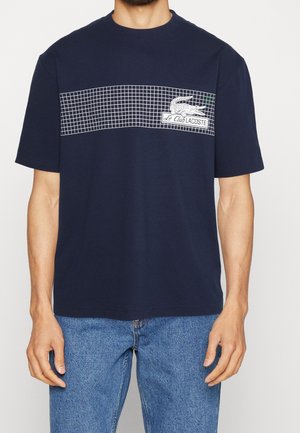 Mężczyzna w granatowym t-shircie z białym wzorem w kratę i logo „Le Club Lacoste” z krokodylem na piersi, w zestawieniu z niebieskimi dżinsami.