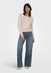 Pull en tricot rose clair avec un col large, associé à un jean bleu à jambes larges et des talons crème. Texture lisse et coupe décontractée.