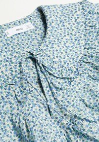 Bluse mit floralem Muster in Blau und Grün, hergestellt aus leichtem Stoff. Verfügt über Rüschen entlang des Kragens und der Knopfleiste.