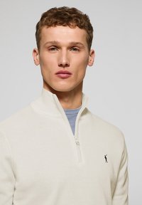 Suéter crema con cuello de cremallera y textura suave; presenta una camiseta interior azul visible debajo; acento con logo en el lado izquierdo del pecho.
