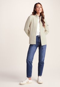 Light green fleece jacket with a high collar, zippered front, and side pockets, paired with blue jeans and white sneakers.
