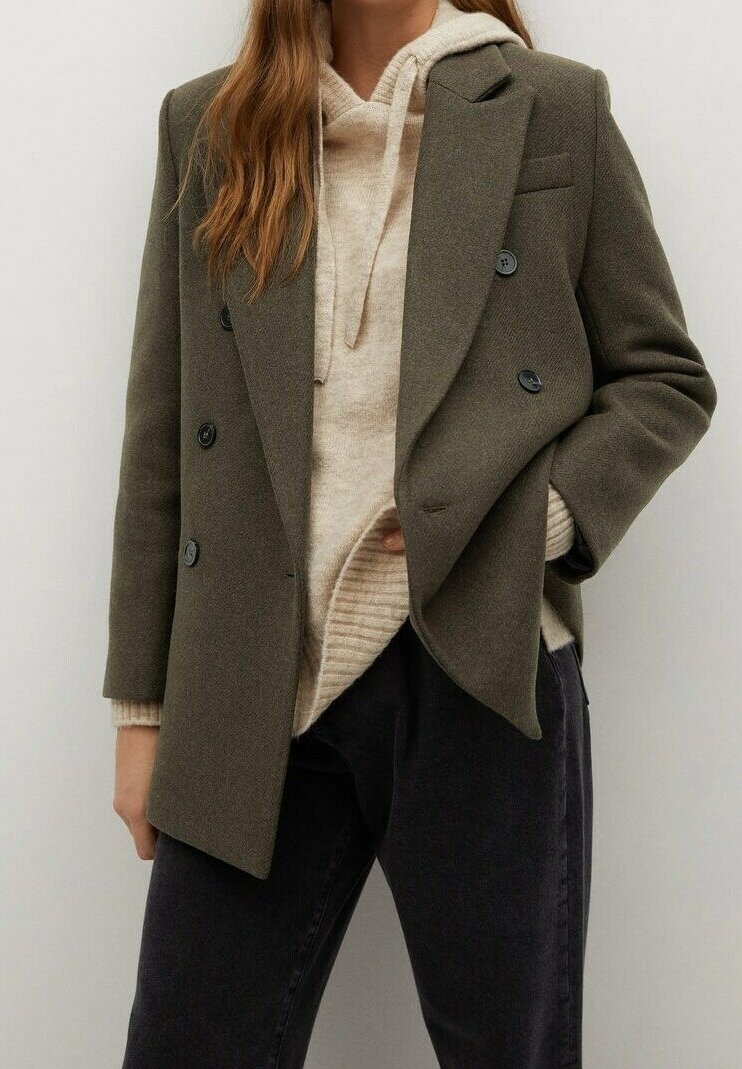 Olivově zelený dvouřadý kabát vyrobený z texturované látky, s černými knoflíky. Nosený přes béžovou mikinu a kalhoty tmavé barvy.