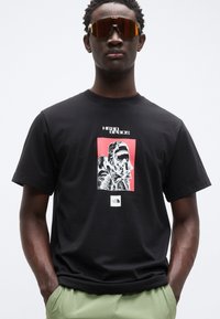 Μαύρο βαμβακερό t-shirt με γραφικό εκτύπωση ενός ατόμου με κουκούλα πάνω σε κόκκινο φόντο, με το λογότυπο The North Face στο στρίφωμα.