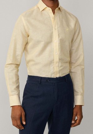 Homme portant une chemise à rayures jaune clair boutonnée, rentrée dans un pantalon bleu marine foncé, debout devant un fond uni.