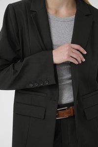 Fekete, szabott blézer négy gombos mandzsettákkal, két elülső zsebbel és nyakkal, szürke bordázott felső és barna bőr öv fölött viselve.