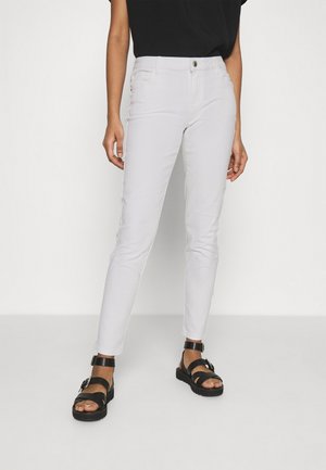 Personne portant un pantalon blanc ajusté, des sandales noires à bride à la cheville et un haut noir, debout devant un fond clair uni.