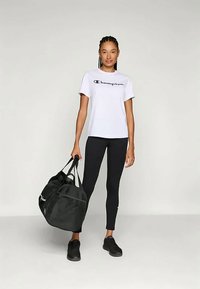 Hvid t-shirt med "Champion" logo, sorte leggings, sort sportstaske og sorte sneakers. Modell står oprejst og viser et afslappet atletisk look.