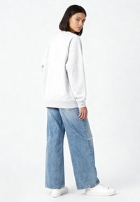 Felpa grigio chiaro con polsini e orlo a coste, abbinata a jeans larghi in denim azzurro chiaro. Modello indossa sneakers bianche, vista da dietro.