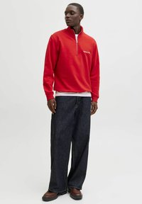 Felpa rossa con zip e collo alto, indossata sopra una camicia bianca, abbinata a pantaloni di denim scuri e larghi e scarpe marroni.