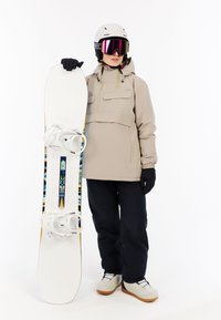 Snowboarder in beiger Jacke mit zwei Brusttaschen, schwarzen Hosen und einem weißen Snowboard mit bunten Mustern und transparenten Bindungen.
