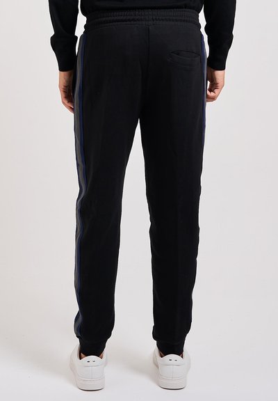 Hombre vestido con pantalones joggers negros con franjas laterales azules y grises y zapatillas blancas, de pie contra un fondo claro y liso.