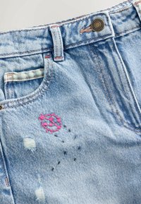 Svetlo modre jeans hlače iz denima z rožnato vezeno pikapolonico in črno črtasto vezenino blizu sprednjega žepa ter obrabljenimi krpami na tkanini.