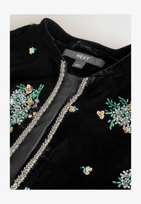 Chaqueta de terciopelo negro con un adorno decorativo de cuentas plateadas y lentejuelas a lo largo del frente, con acentos en verde y dorado.