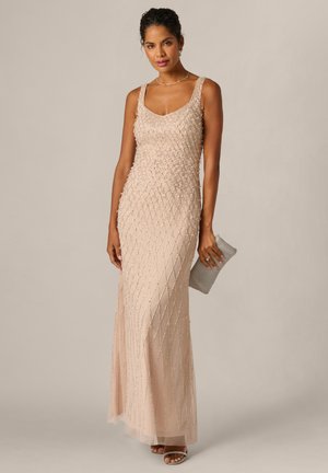 Kvinde iført en ærmeløs, gulvlængde beige aftenkjole med perlebesætning, der holder en tekstureret clutch-taske, stående på en ensfarvet baggrund.