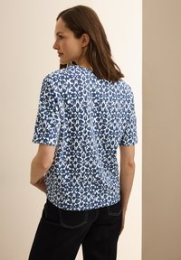 Halenka s krátkým rukávem v námořnické modré barvě s bílým vzorem, která má abstraktní design. Materiál vypadá měkce, s uvolněným střihem a zaobleným lemem.