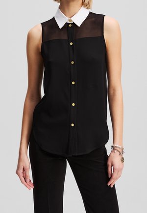 Mouwloze zwarte blouse met doorzichtige bovenkant, witte puntige kraag en gouden knopen, gedragen met een zwarte broek en bedelarmbanden.