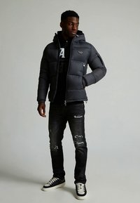 Tumši pelēka iesaiņojoša jaka ar kapuci, uz krūtīm ar logo, kombinēta ar melnām nodilušām džinsa biksēm un melniem sporta apaviem ar baltām zolēm.