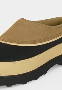 Κοντινή λήψη ενός παπουτσιού slip-on με καφέ δερμάτινο επάνω μέρος, μαύρο μεσαίο τμήμα από καουτσούκ, μπεζ λεπτομέρεια και μαύρη υφή στην σόλα.
