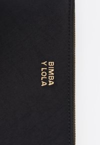 Bolsa textil negra con una superficie lisa y el logo de marca en relieve dorado "Bimba y Lola" en el frente. Detalle de cremallera visible.