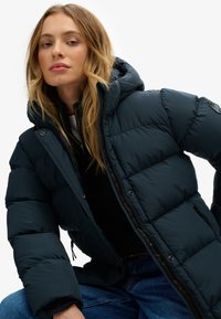 Giacca imbottita blu scuro con cappuccio, caratterizzata da una texture trapuntata, chiusura con zip e tasche laterali; indossata sopra una maglia nera e jeans blu.