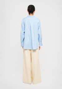 Light blue and white striped oversized shirt with a back pleat and a collar, paired with beige wide-leg trousers. Smooth fabric texture.