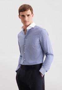 Camicia a righe blu e bianca. Presenta un colletto button-down, taschino frontale e maniche lunghe con polsini abbottonati. Vestibilità tailored.