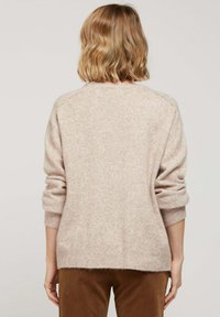 Un pull tricoté rose clair avec une texture douce, featuring des épaules tombantes et des poignets côtelés, porté sur un pantalon marron.