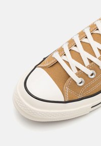 Brun canvas sneaker med vit gummitå, svart kant och vita snören. Texturerat tyg med synliga sömdetaljer.