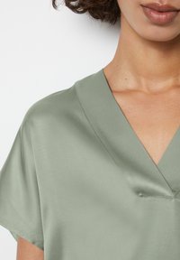 Blusa verde in satin con scollatura a V e maniche corte. Il tessuto ha una texture liscia e una leggera lucentezza. I dettagli delle cuciture sono visibili vicino alla scollatura.