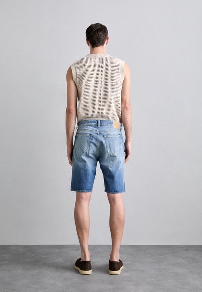 Shorts din denim de culoare albastru deschis cu un aspect decolorat, asortați cu un top fără mâneci tricotat de culoare bej. Modelul stă pe un teren gri, în fața unui fundal simplu.